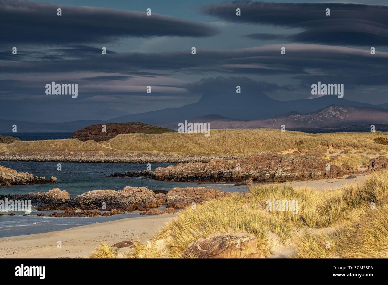 A small sandy beach with dune grass. Mountains in the background and exciting clouds in the sky. Stock Photo