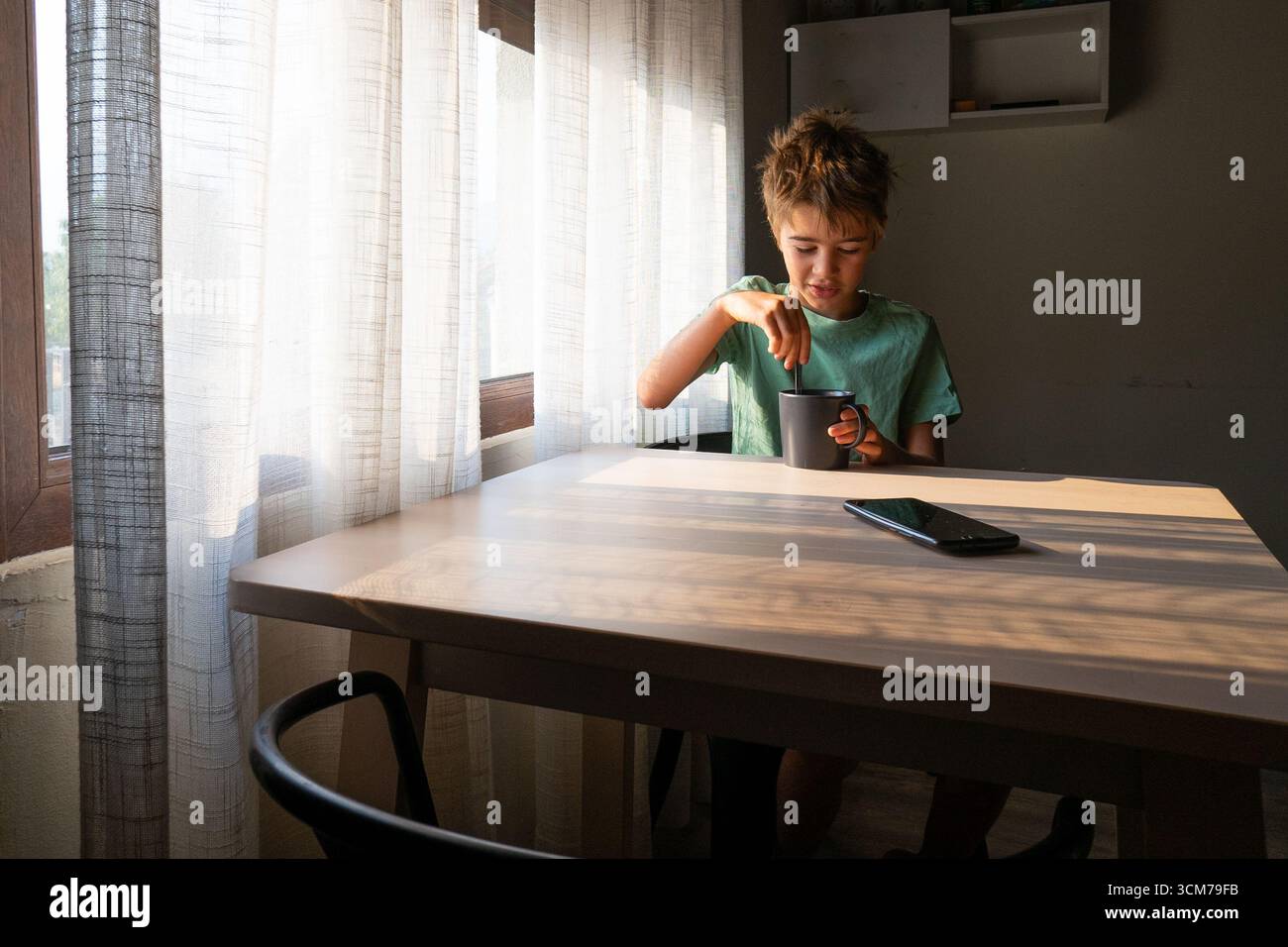 Child stirring a warm beverage at a wooden table in a sunlit room, with a smartphone resting beside the mug Stock Photo