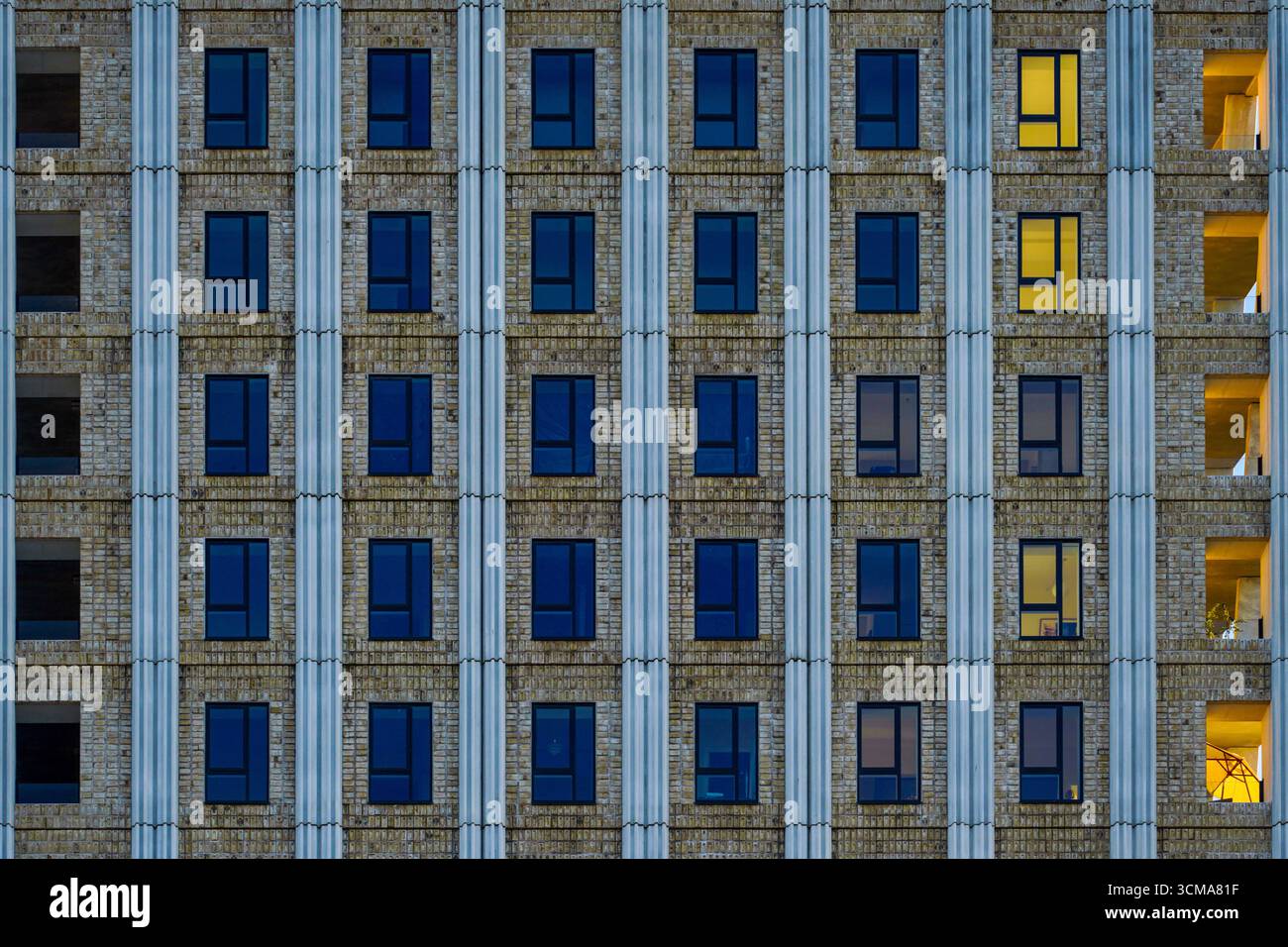 Playful Harmony: Verticality and Illumination. A night view of a building facade in Copenhagen, Denmark shows earth-toned masonry combined with vertical elements and evenly arranged windows, where alternating patterns of dark and illuminated panes create a rhythmic architectural composition. Stock Photo