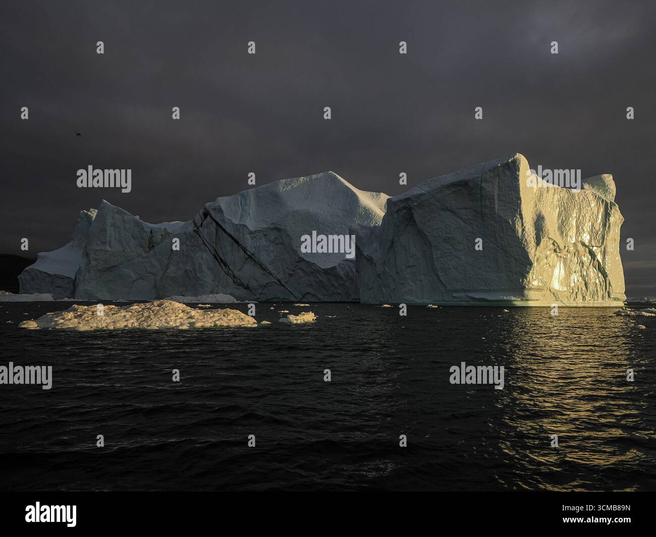 Large iceberg illuminated by sunset light under dark clouds in ocean near Disko Bay, Greenland Stock Photo