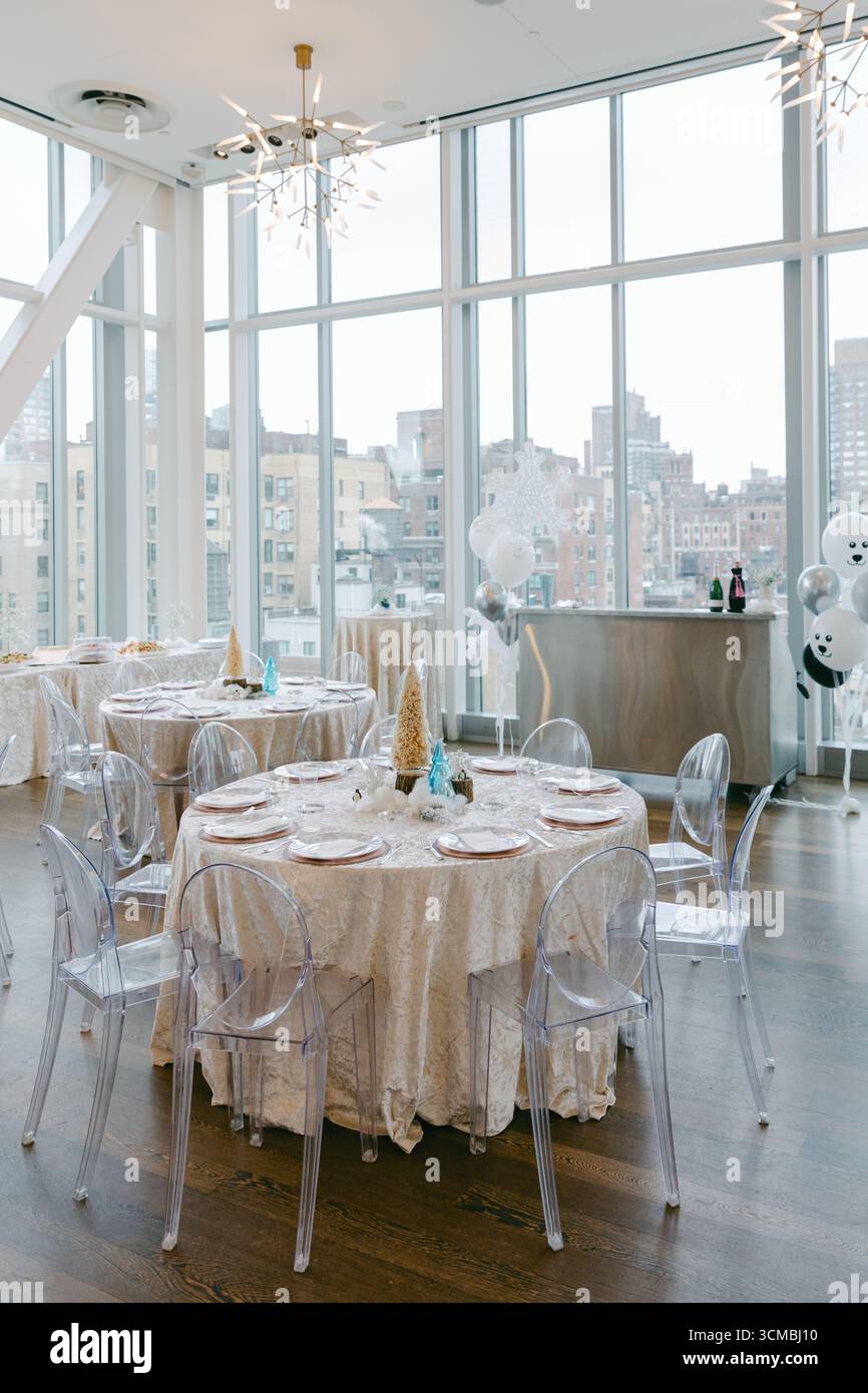 A bright event space features round tables with white tablecloths and transparent chairs, providing a city skyline view. Stock Photo