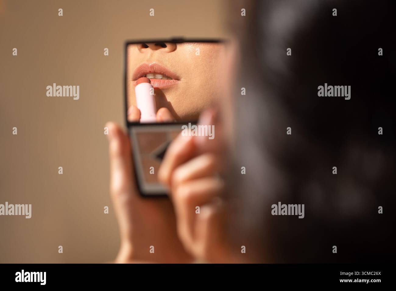 Female individual is applying lipstick while looking into a handheld mirror, highlighting beauty routine and self-care in a softly lit environment Stock Photo