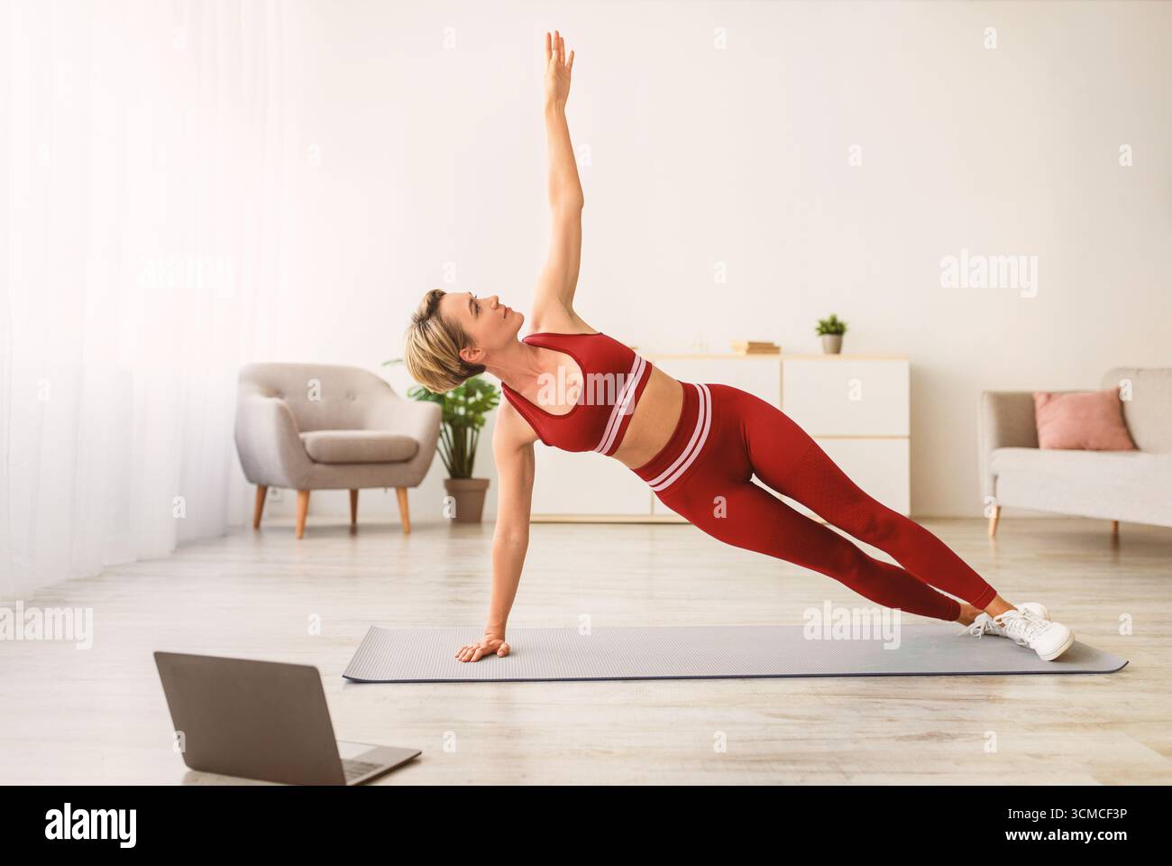 Middle-aged woman doing side plank on mat with pc Stock Photo
