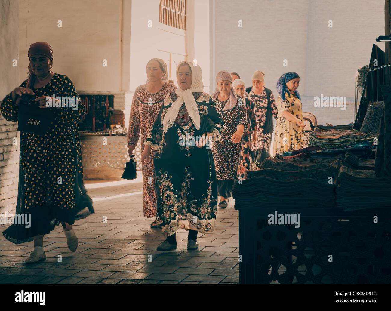 Group of Uzbek women in patterned dresses and headscarves browse textiles at sunlit market stall, Bukhara, Uzbekistan. Stock Photo