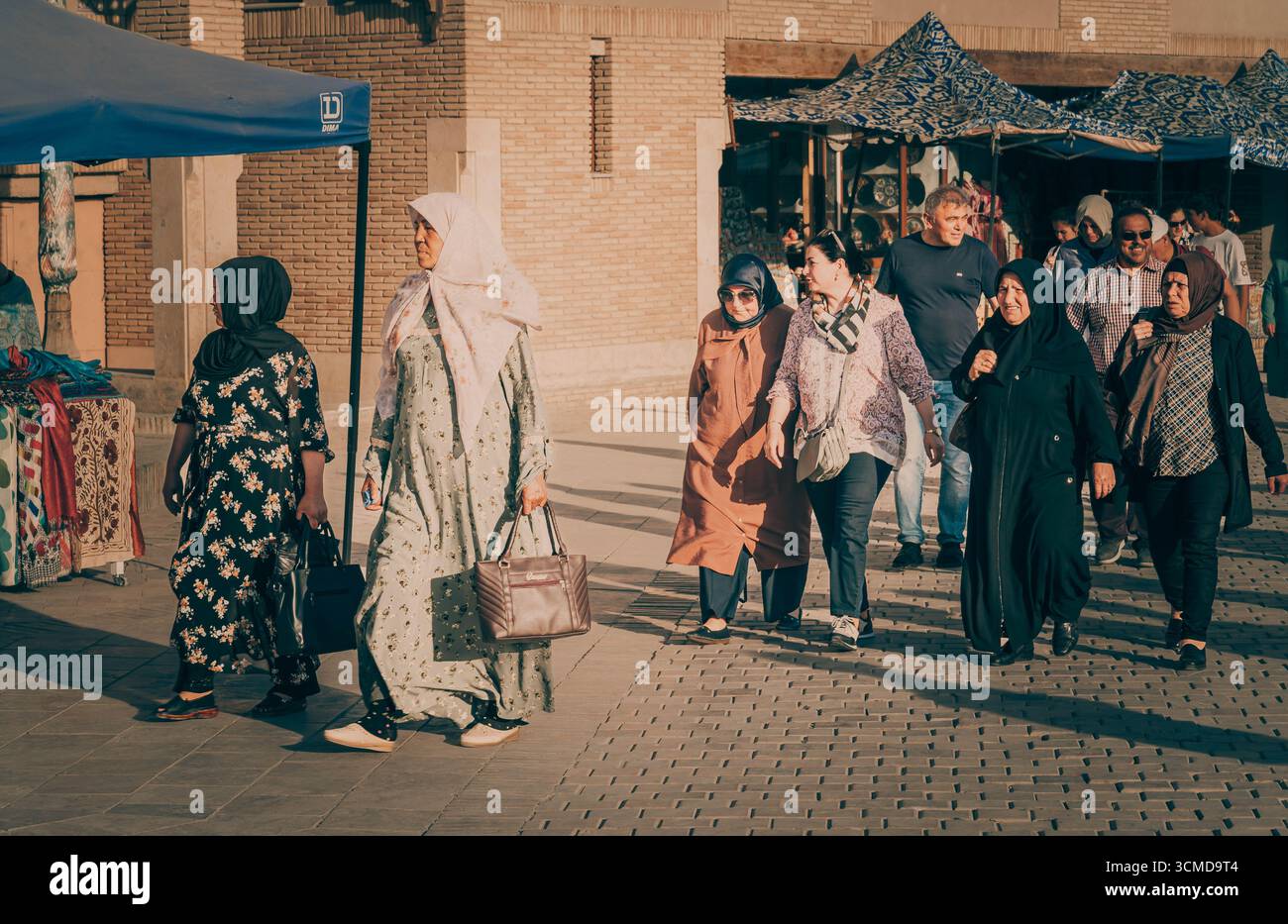 Group of Uzbek women and men in traditional and modern attire stroll past sunlit market tents, Bukhara, Uzbekistan, with vibrant textiles and handmade Stock Photo