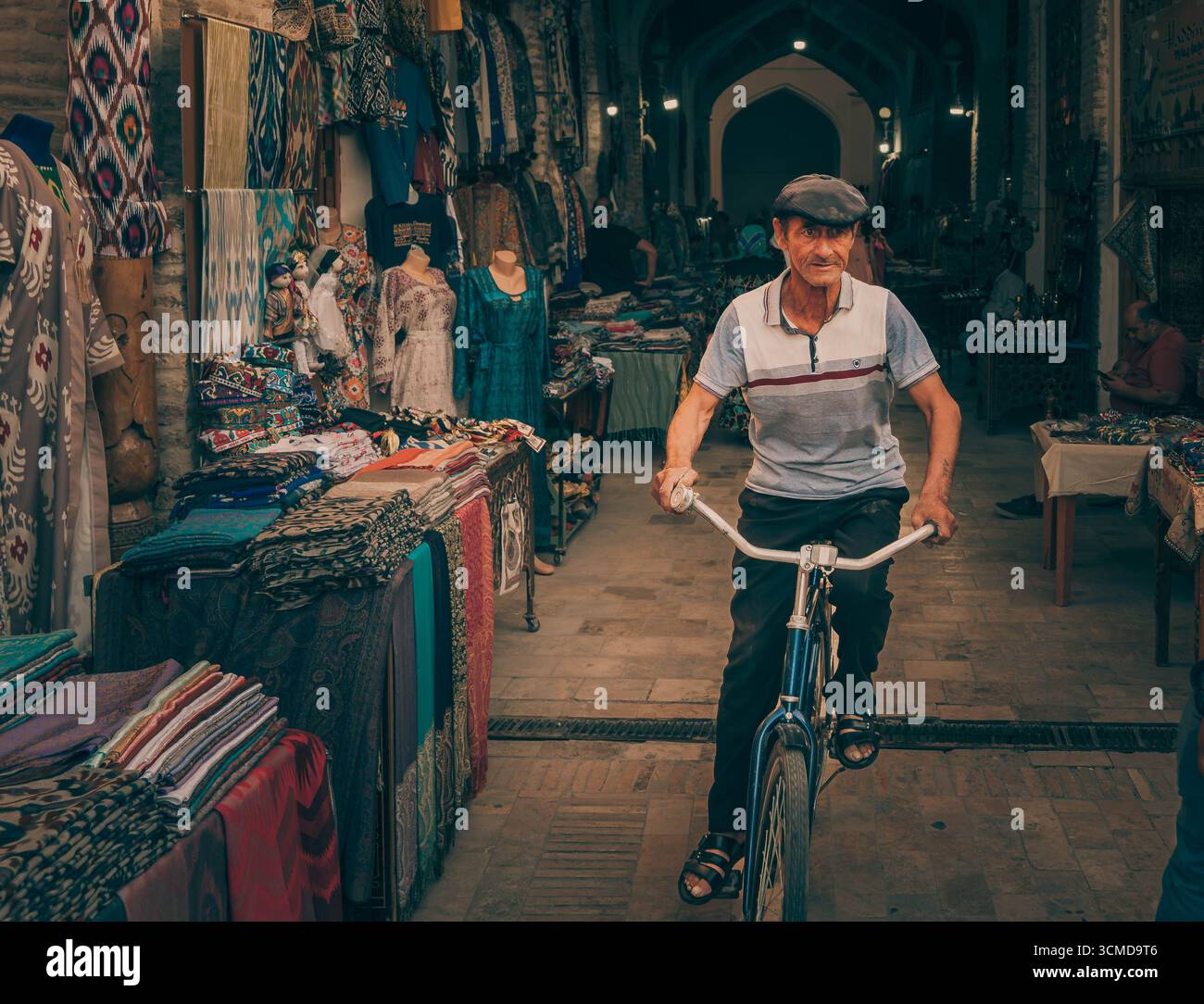 Elderly Uzbek man cycling through bustling bazaar, Bukhara, Uzbekistan, amid colorful textiles, embodying vibrant market life in trading hub. Stock Photo