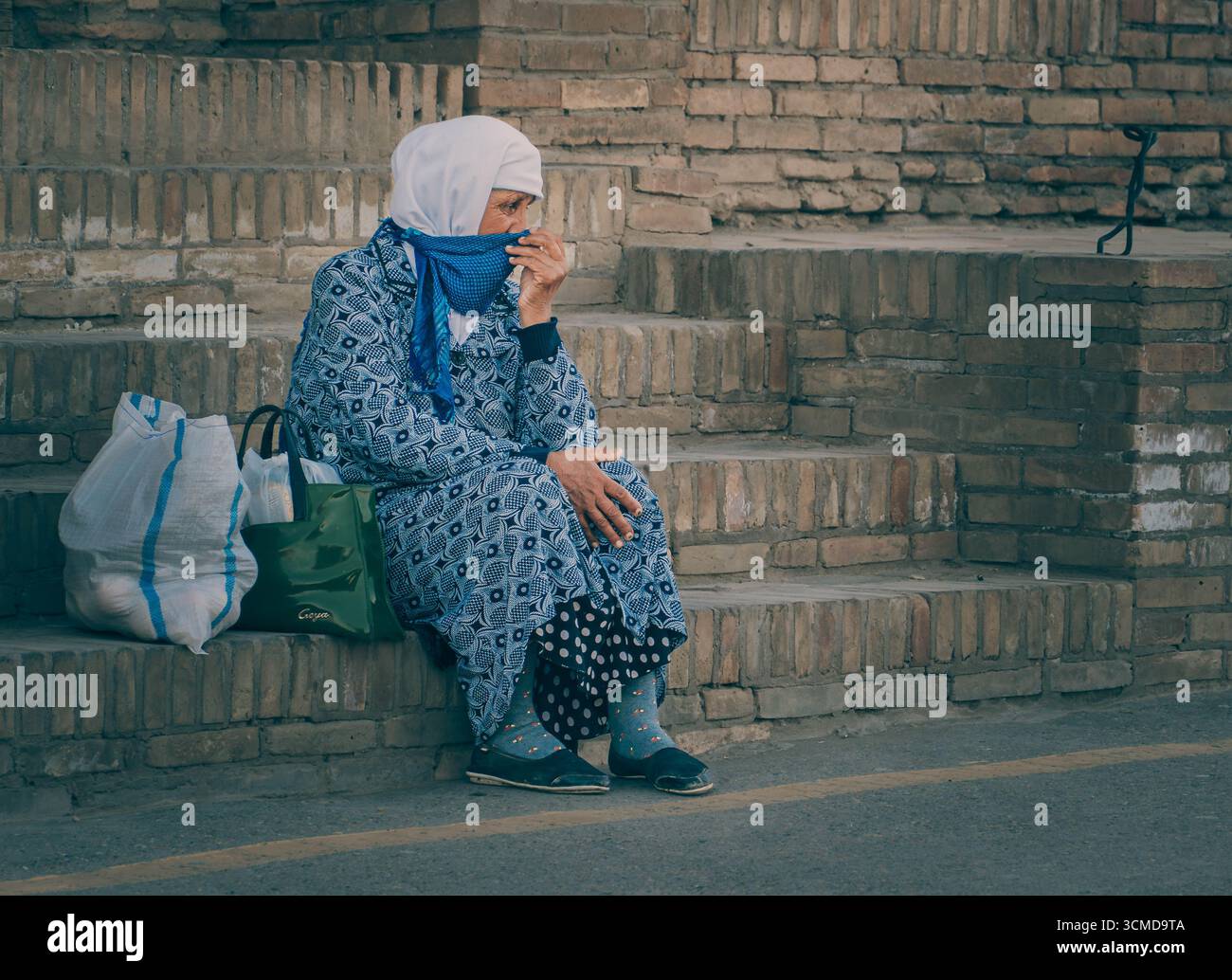 Elderly Uzbek woman sits on brick steps in Bukhara, Uzbekistan, adjusting blue scarf over face, wearing patterned blue dress. Stock Photo