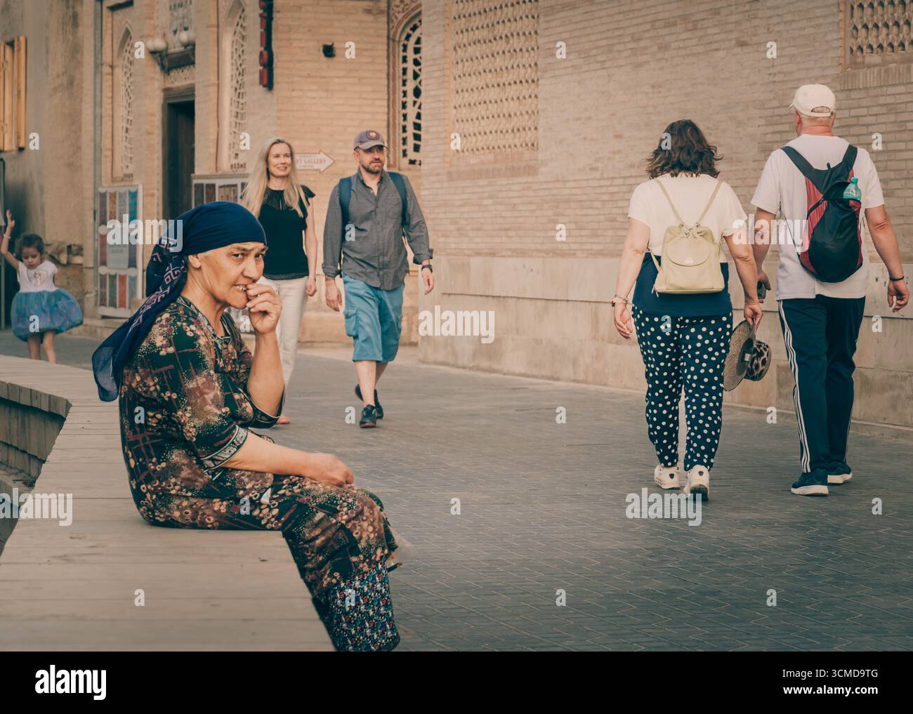 Elderly woman in blue headscarf and patterned dress sits on curb, eating near historic brick buildings, Bukhara, Uzbekistan Stock Photo