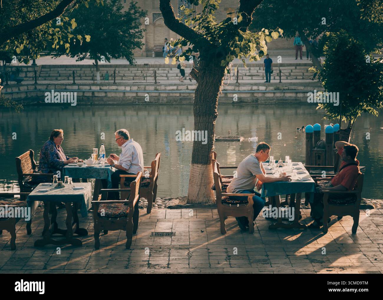 Group of Uzbek adults including elderly and middle-aged people dine at tables by Lyab-i-Hauz pool in Bukhara, Uzbekistan, with food, drinks. Stock Photo