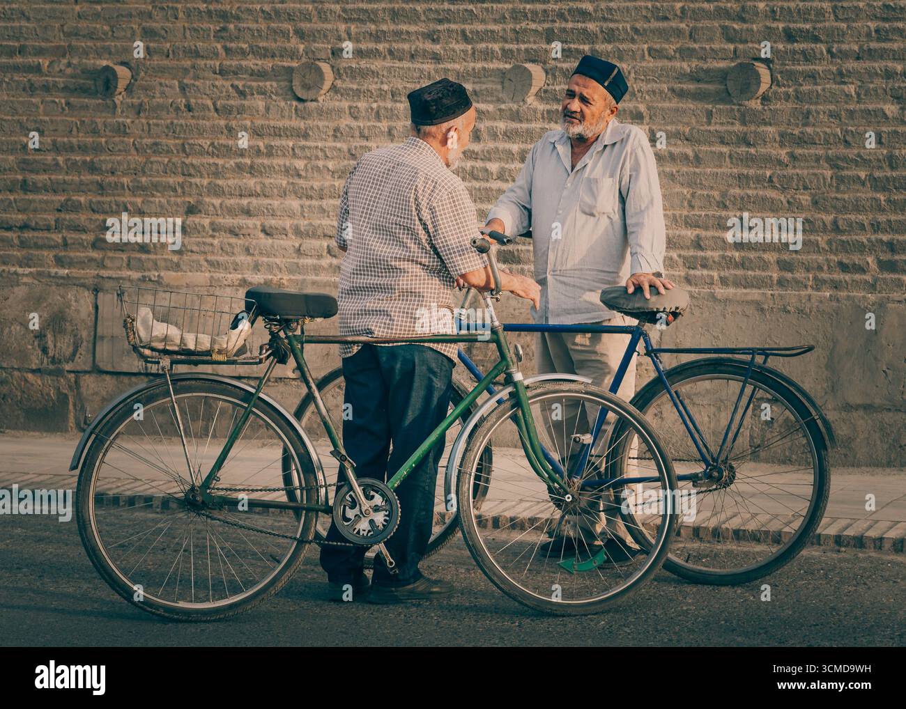Elderly Uzbek men in skullcaps chatting, Bukhara, Uzbekistan, leaning on bicycles, showcasing community ties in historic Central Asian setting. Stock Photo