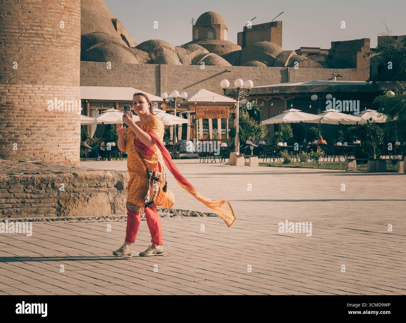 Young Uzbek woman walks and takes a photo with her phone in Bukhara, Uzbekistan, wearing a vibrant orange outfit with a flowing scarf. Stock Photo