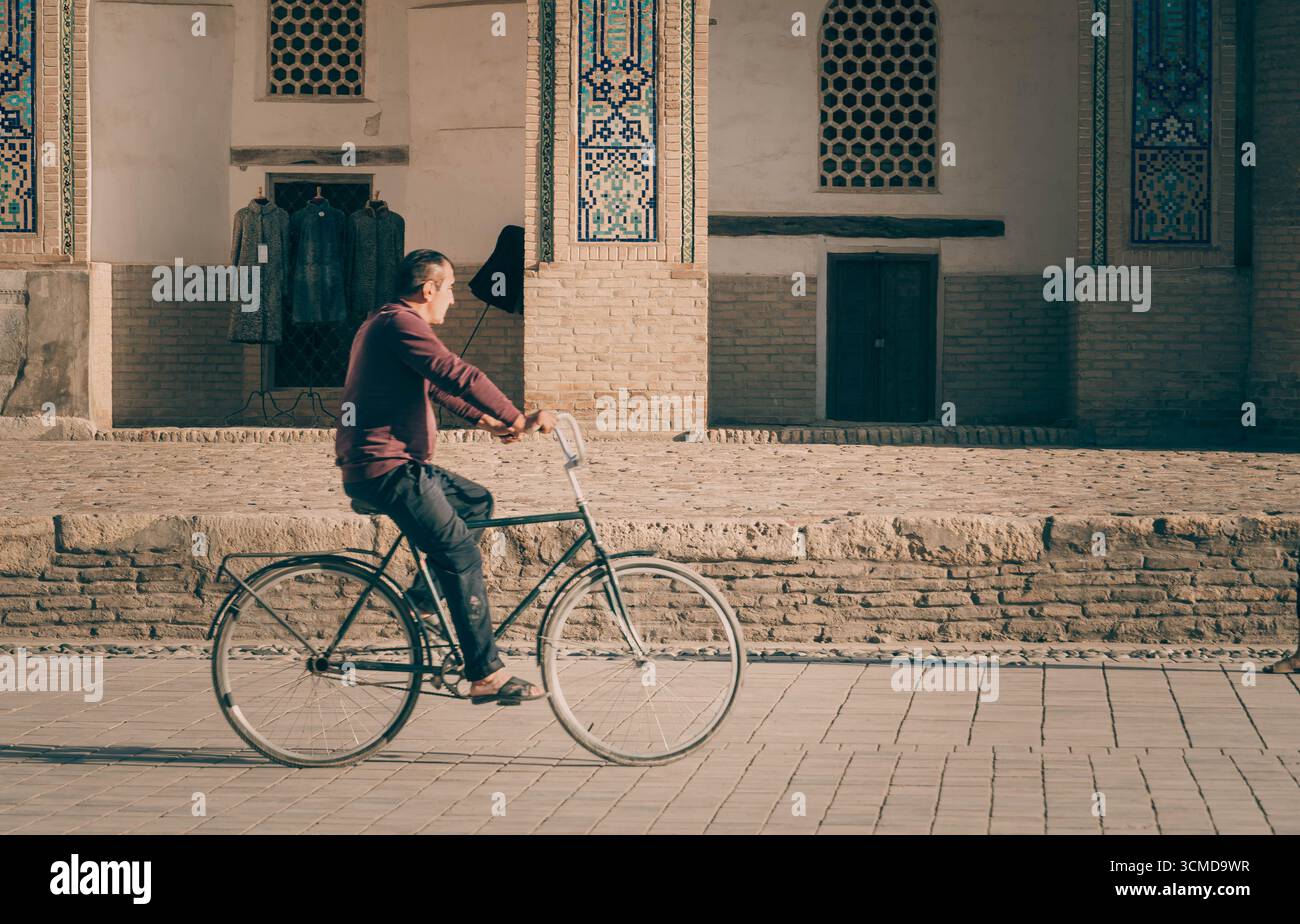 Middle-aged Uzbek man riding bicycle, Bukhara, Uzbekistan, in alley between brick walls, reflecting local transport in Silk Road city. Stock Photo