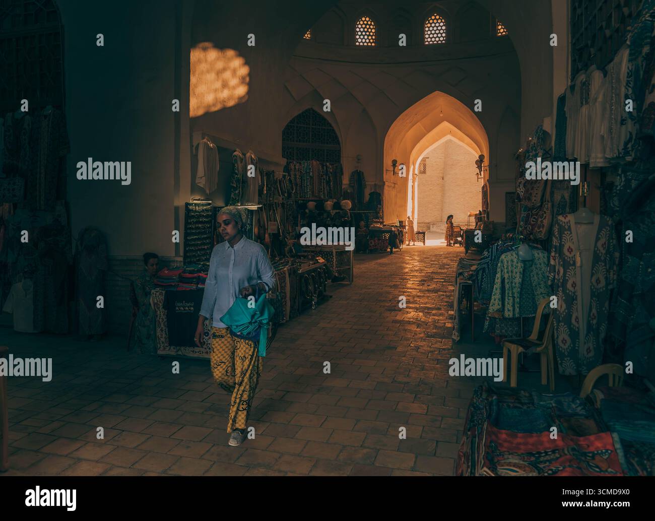 Young Uzbek woman walks through an indoor bazaar in Bukhara, Uzbekistan, amid hanging textiles and arched hallways, wearing headscarf. Stock Photo