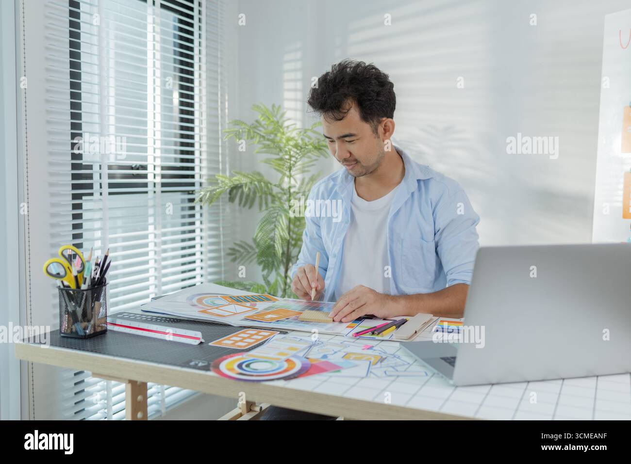 A focused designer is working on UX/UI design ideas, analyzing color palettes and interface structures pinned on a board. The scene reflects creativit Stock Photo