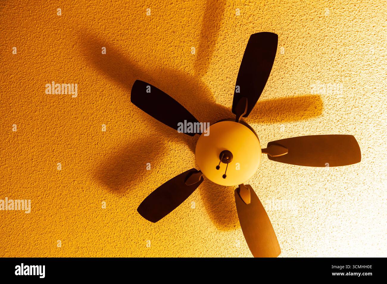 Ceiling fan casting a rotating shadow on the ceiling surface with soft yellow light, close up Stock Photo