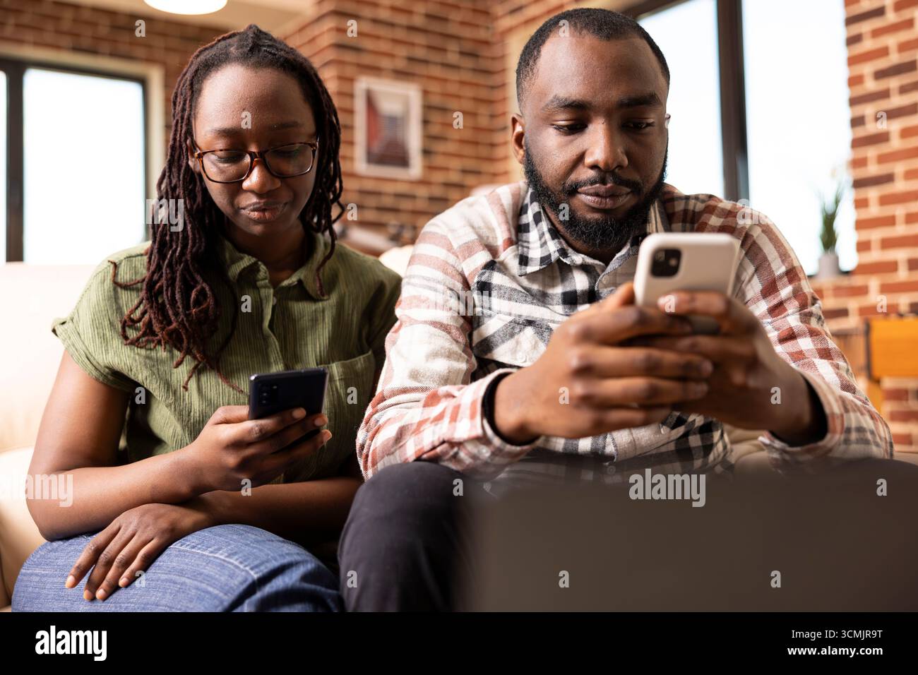 African american husband and wife sitting on couch, each focused on ...