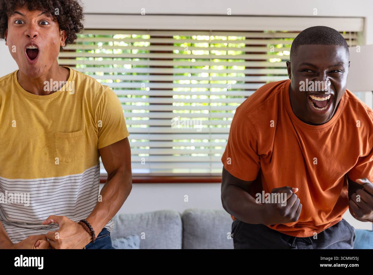 Diverse male friends cheering and leaning forward in living room near couch with decorative pillows Stock Photo