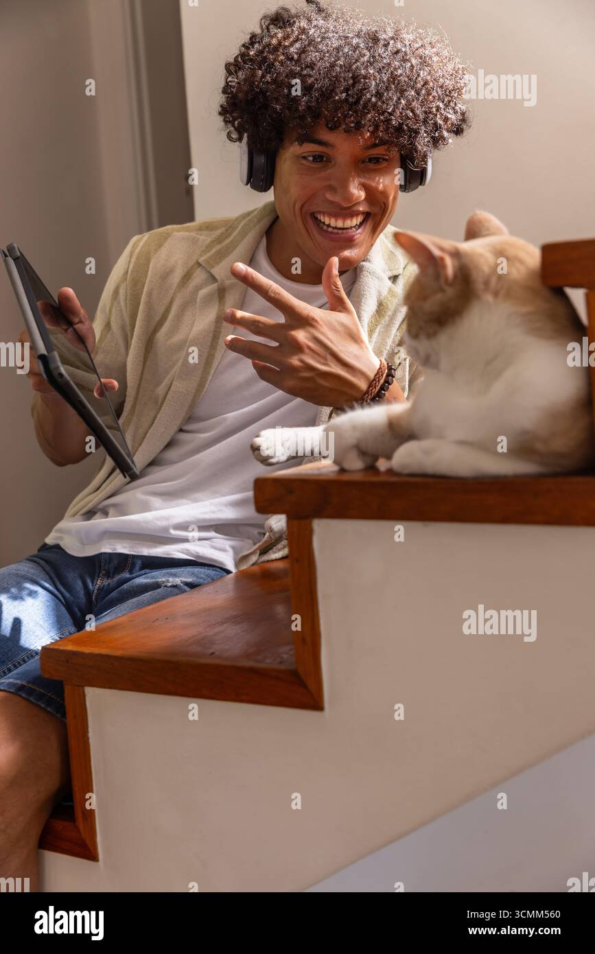 Man sitting on staircase using tablet and headphones while cat basking in sunlight Stock Photo