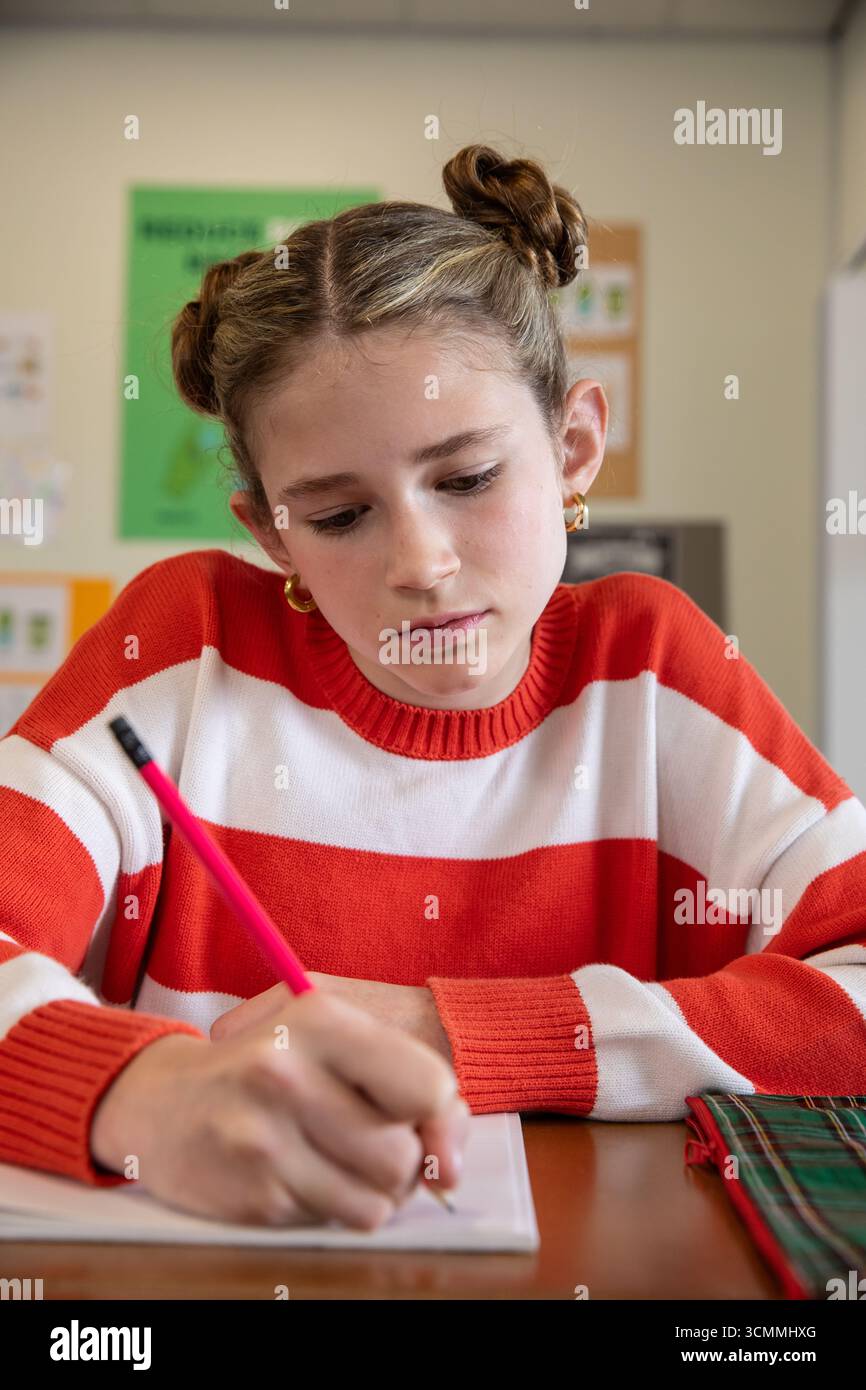 Girl wearing red sweater writing in notebook with pink pencil, plaid case on classroom desk Stock Photo