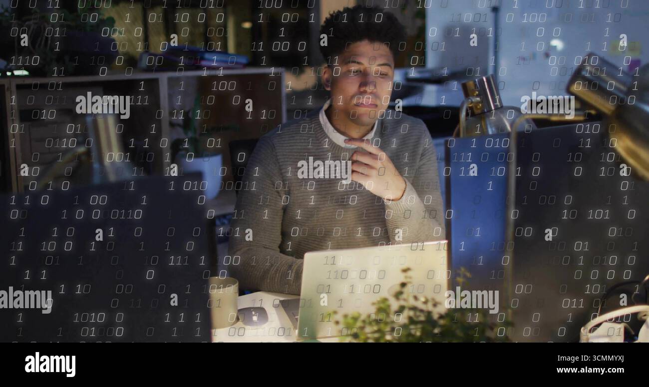 Developer wearing grey sweater analyzing binary code in office at night, with laptop and desk lamp Stock Photo