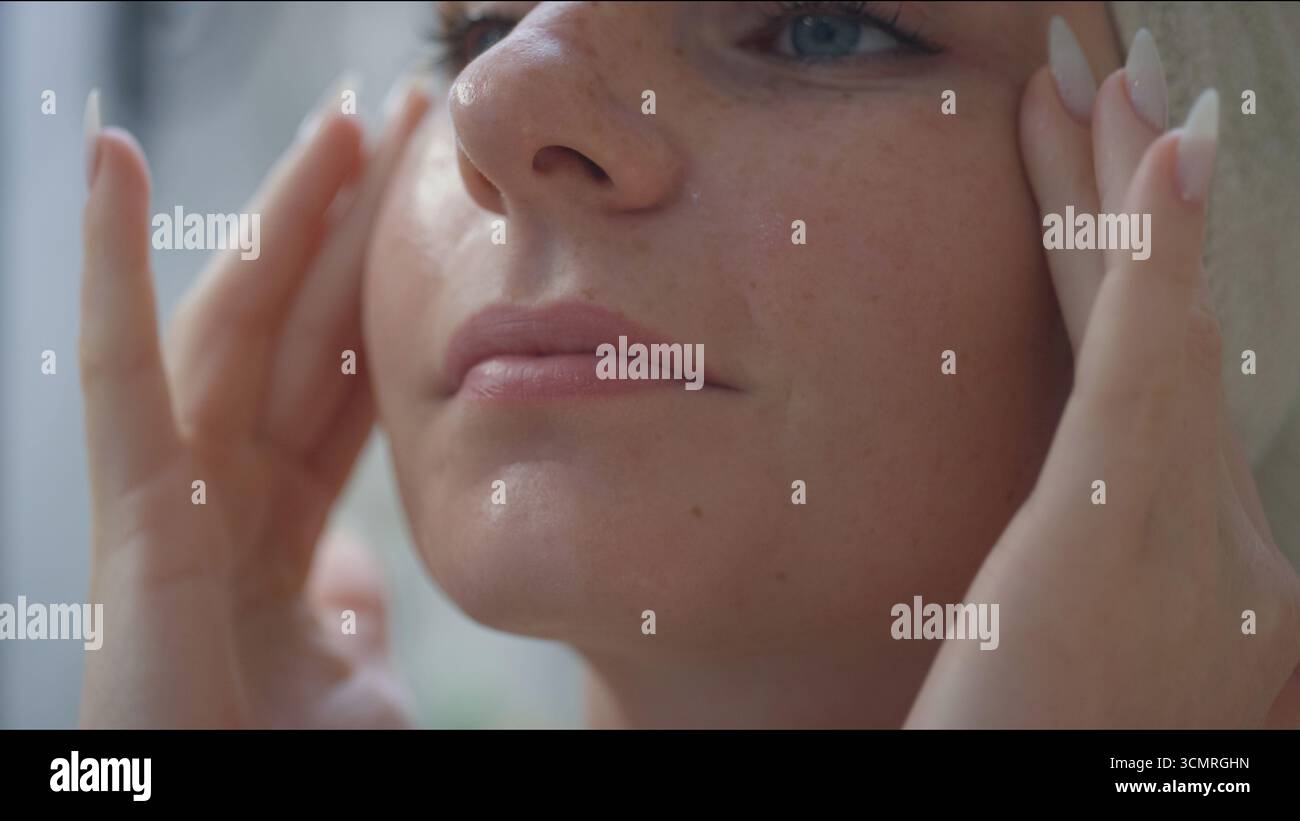 A young woman gently massages her face while applying a serum in a bright bathroom, focusing on her skincare regimen. Stock Photo