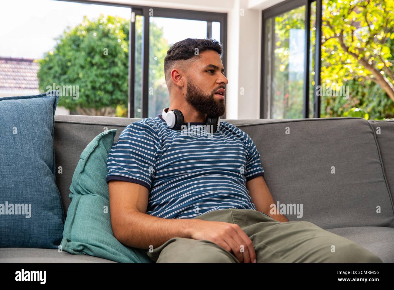 Man wearing striped t-shirt sitting on sofa at home with windows, teal cushions and headphones Stock Photo