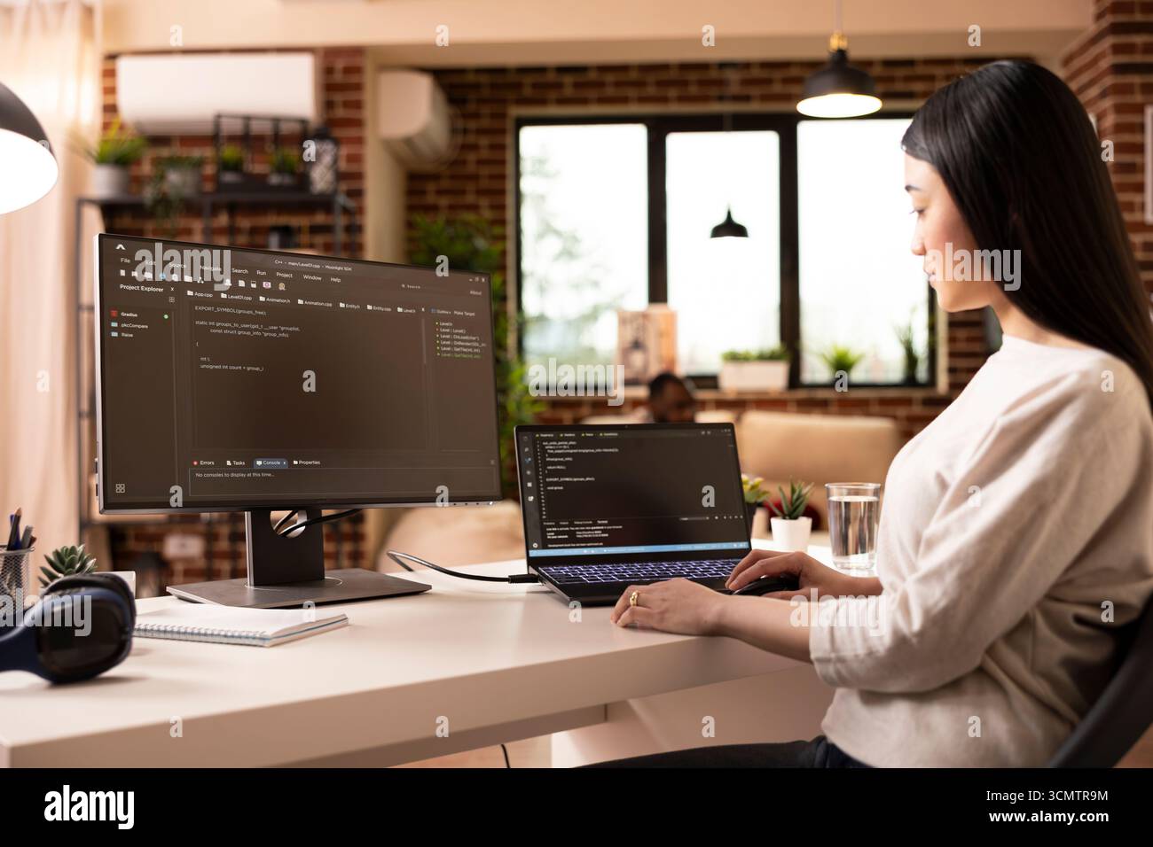 Focused female developer programming on laptop in home office. Asian woman building software and coding website remotely from modern apartment using digital tools for web development. Stock Photo