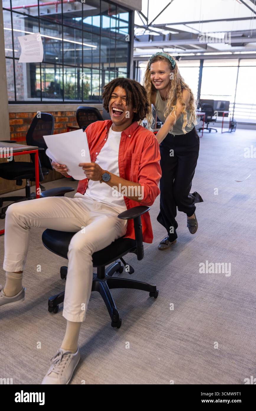 Diverse coworkers pushing black swivel chair down office corridor with ...