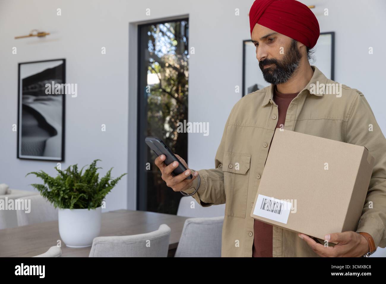 Indian man standing in dining room holding shipping box, checking smartphone, copy space Stock Photo
