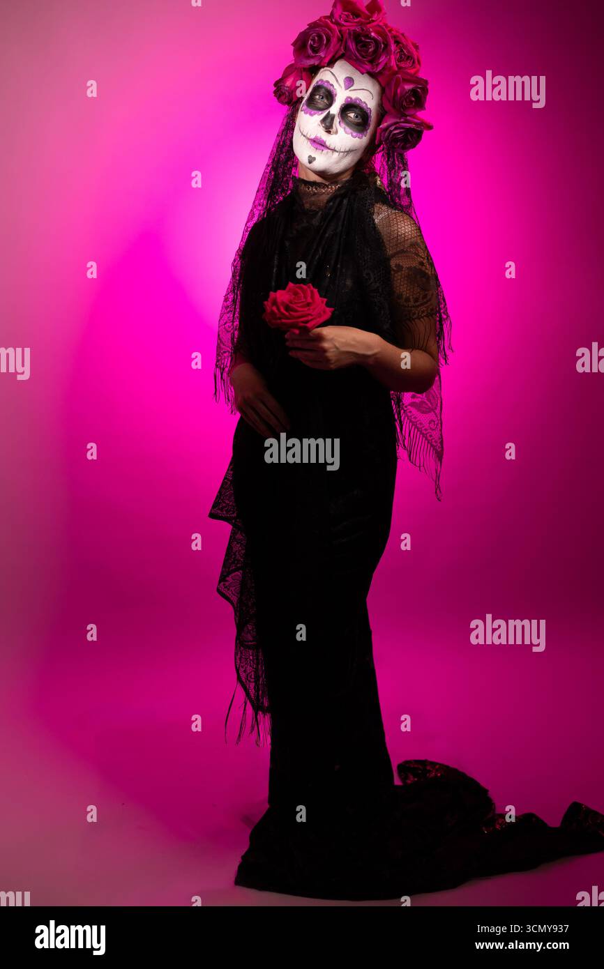 Woman with skull face makeup, wearing a black dress and floral crown, holds a rose. Her costume is for Dia de los Muertos, or Day of the Dead, a Mexican holiday, set against a pink background. Stock Photo