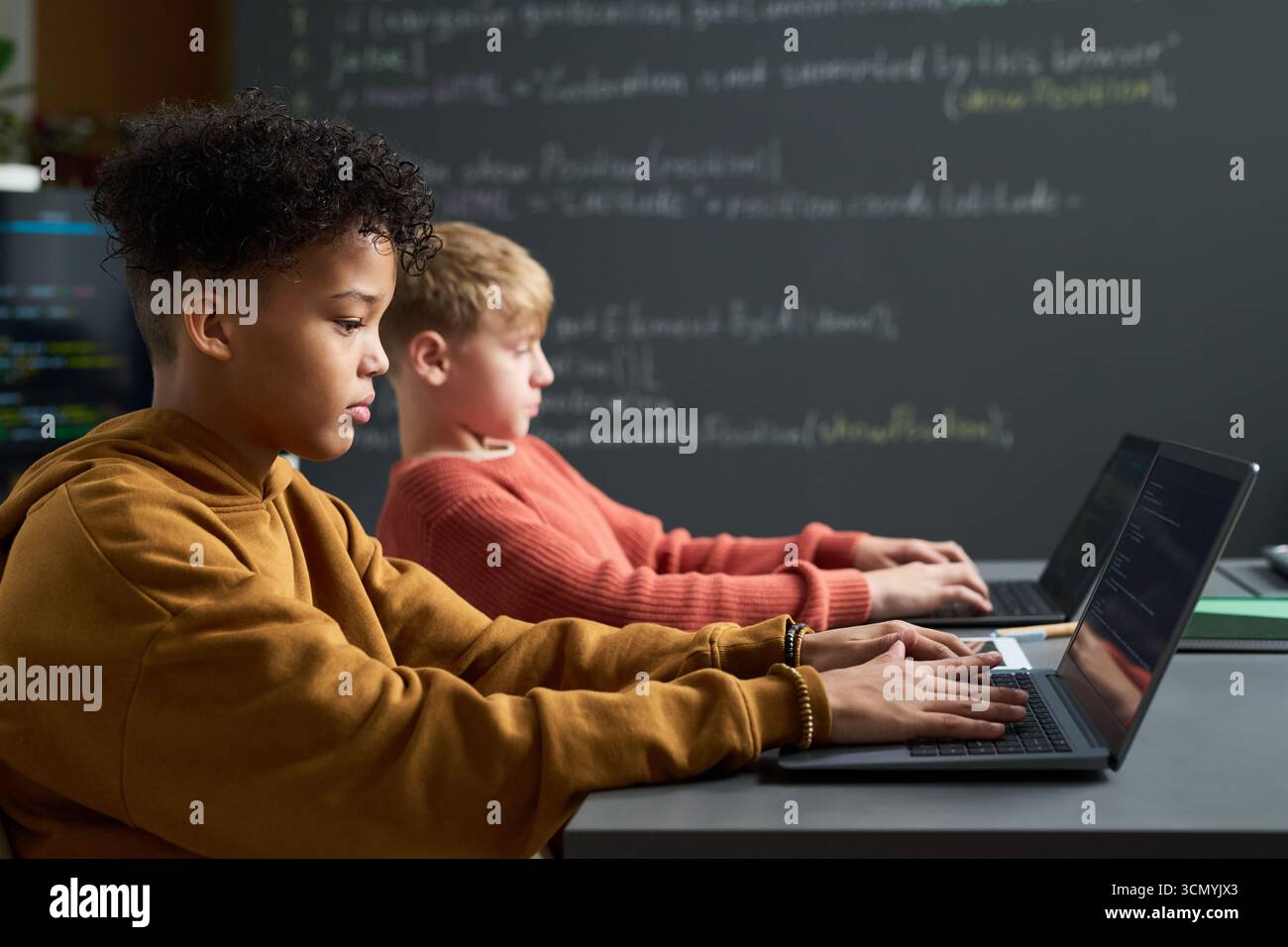 Multiethnic boys sitting side by side using laptops in classroom, focusing on typing and coding, with chalkboard and programming code visible in background, representing technology education Stock Photo