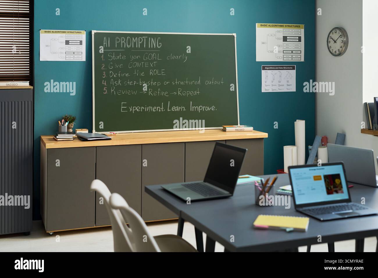 Empty classroom with chalkboard displaying AI prompting instructions, laptops on table in foreground, educational posters on teal wall, clock showing time, sunlight entering through window blinds Stock Photo