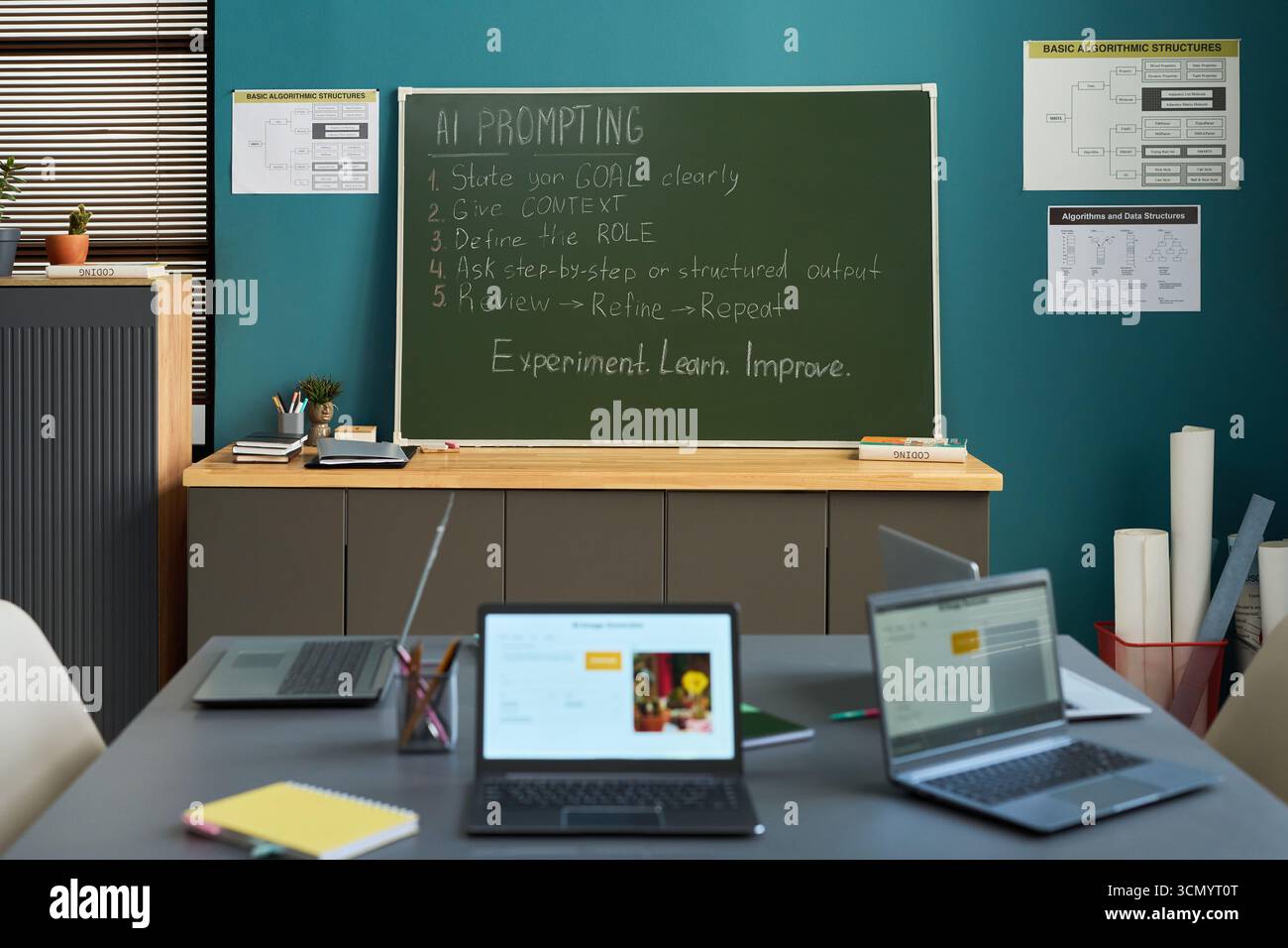 Classroom scene showing empty desks with open laptops in foreground, green chalkboard with AI prompting instructions written in chalk, educational posters on blue wall in background Stock Photo