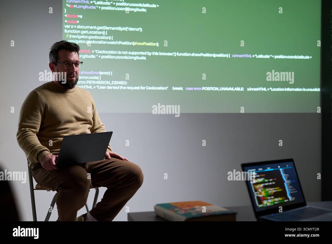 Caucasian man sitting on stool using laptop while presenting programming code projected on wall, holding computer on lap, explaining software development concepts Stock Photo