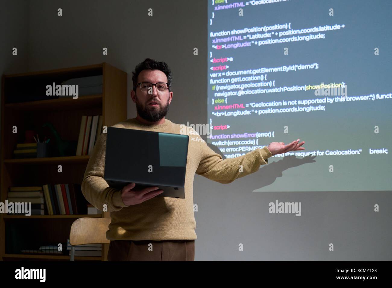 Caucasian young adult man presenting programming code on projection screen while holding laptop, standing in office setting, explaining software development concepts to audience Stock Photo