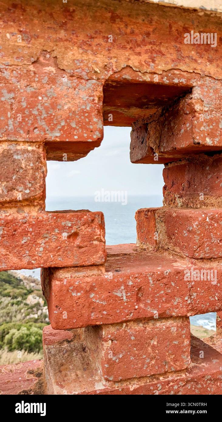 Atlantic Ocean view framed through a cross-shaped opening in weathered red brickwork, contrasting rough textures with soft sky and deep blue sea - Smartphone Captured Stock Image
