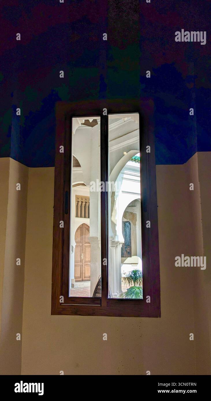 View through wooden-framed window to arched columns, carved door, and courtyard with greenery in bright daylight. - Smartphone Captured Stock Image