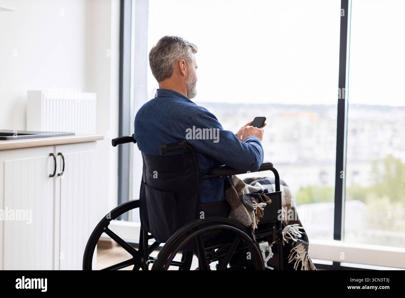 Man in wheelchair using smartphone indoors looking out large window view Stock Photo