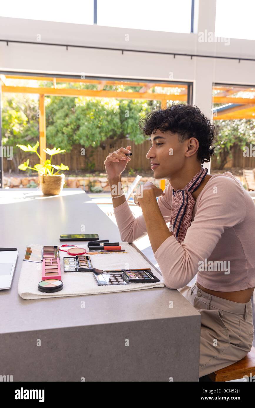 Asian man applying mascara at kitchen island wearing top among makeup palettes brushes phone Stock Photo