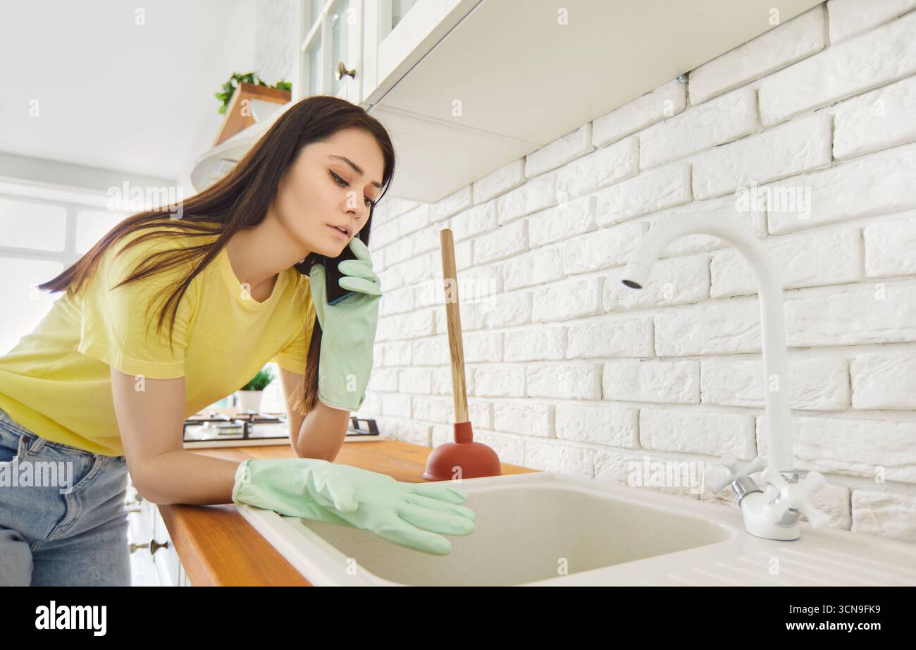 Worried housewoman sing plunger to unclog sink in kitchen using smartphone to call plumbing service Stock Photo