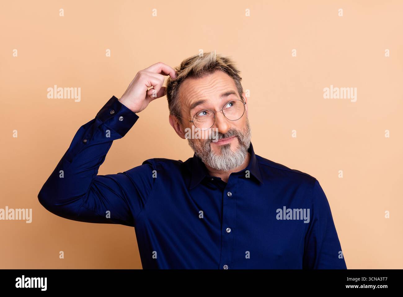 Portrait of a mature businessman in navy blue shirt scratching head with a puzzled expression against beige background Stock Photo