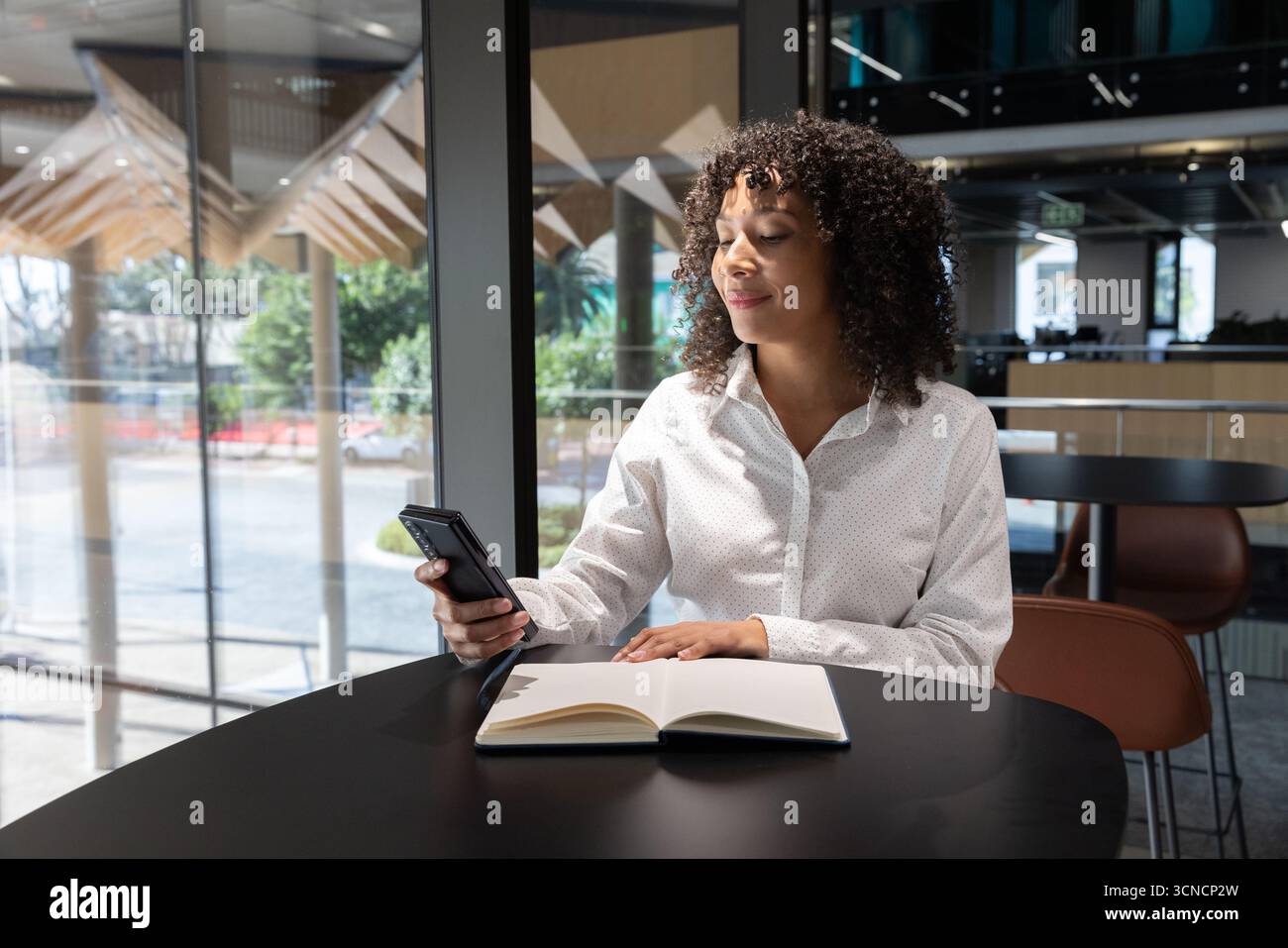 African american woman using smartphone, writing in notebook at lounge table in office Stock Photo
