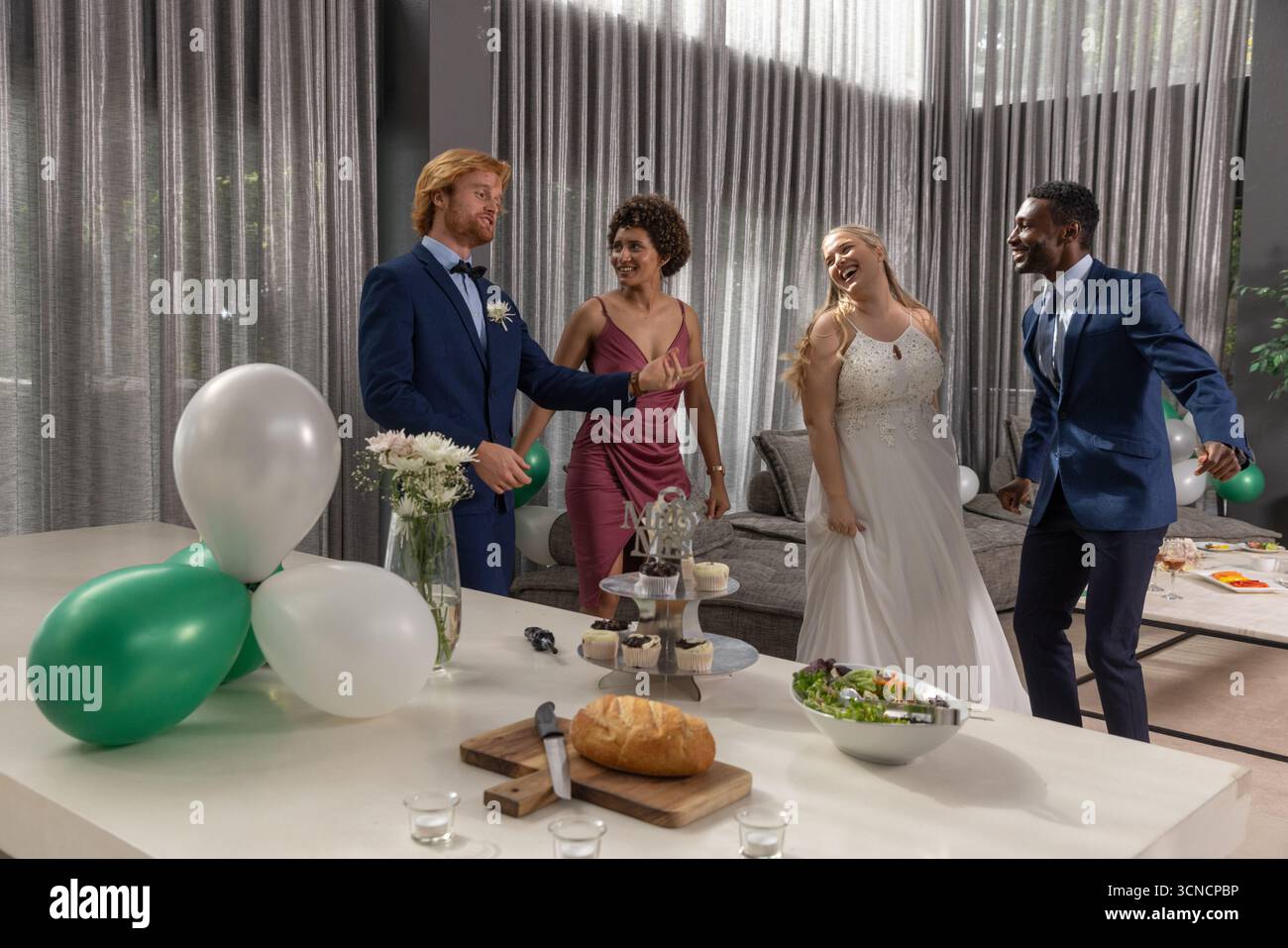 Diverse wedding party dancing and chatting around kitchen island table ...