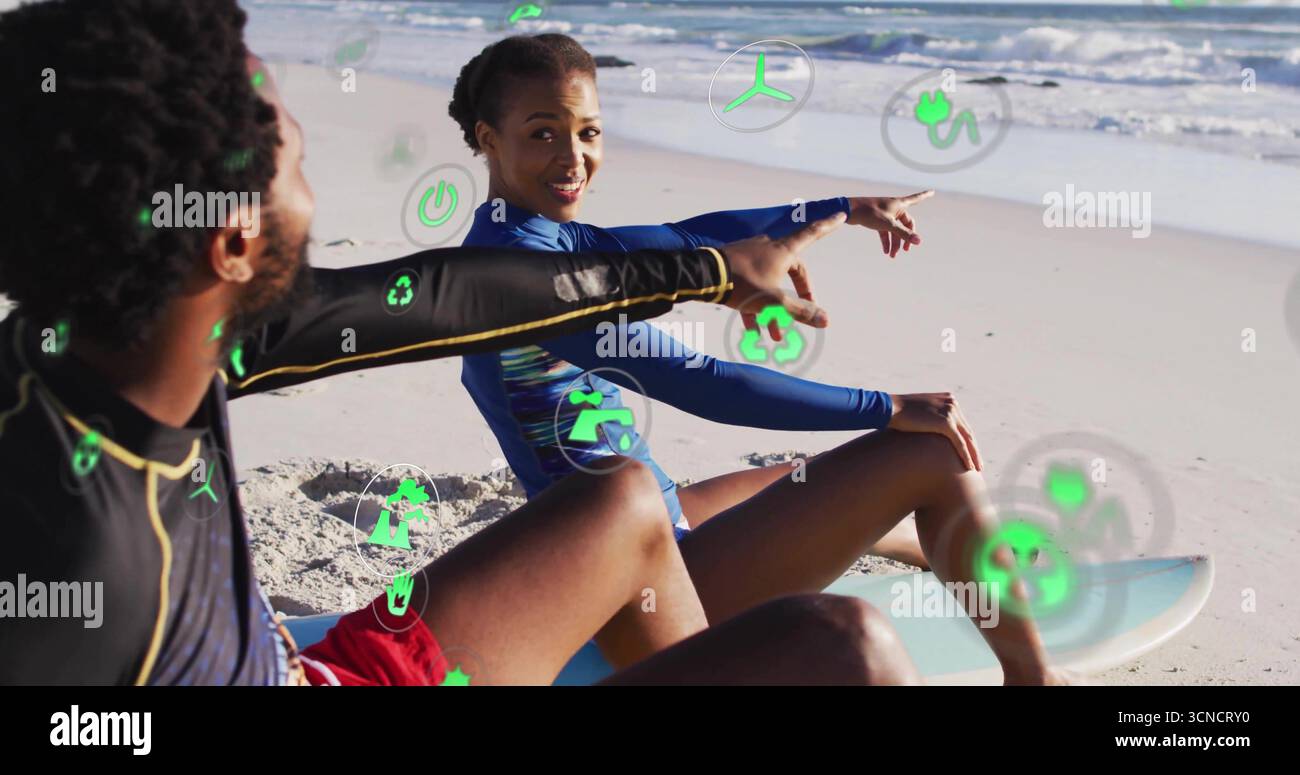 Two surfers in rash guards pointing at waves astride surfboards at shore, with green digital icons Stock Photo