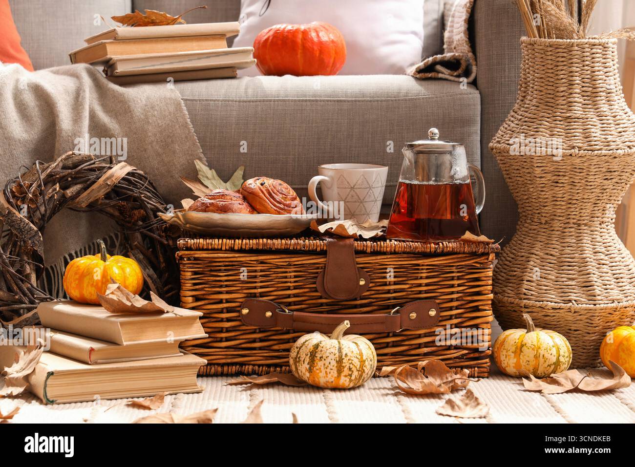 Autumn composition with teapot, cup of hot beverage, tasty buns and pumpkins on wicker basket in living room Stock Photo
