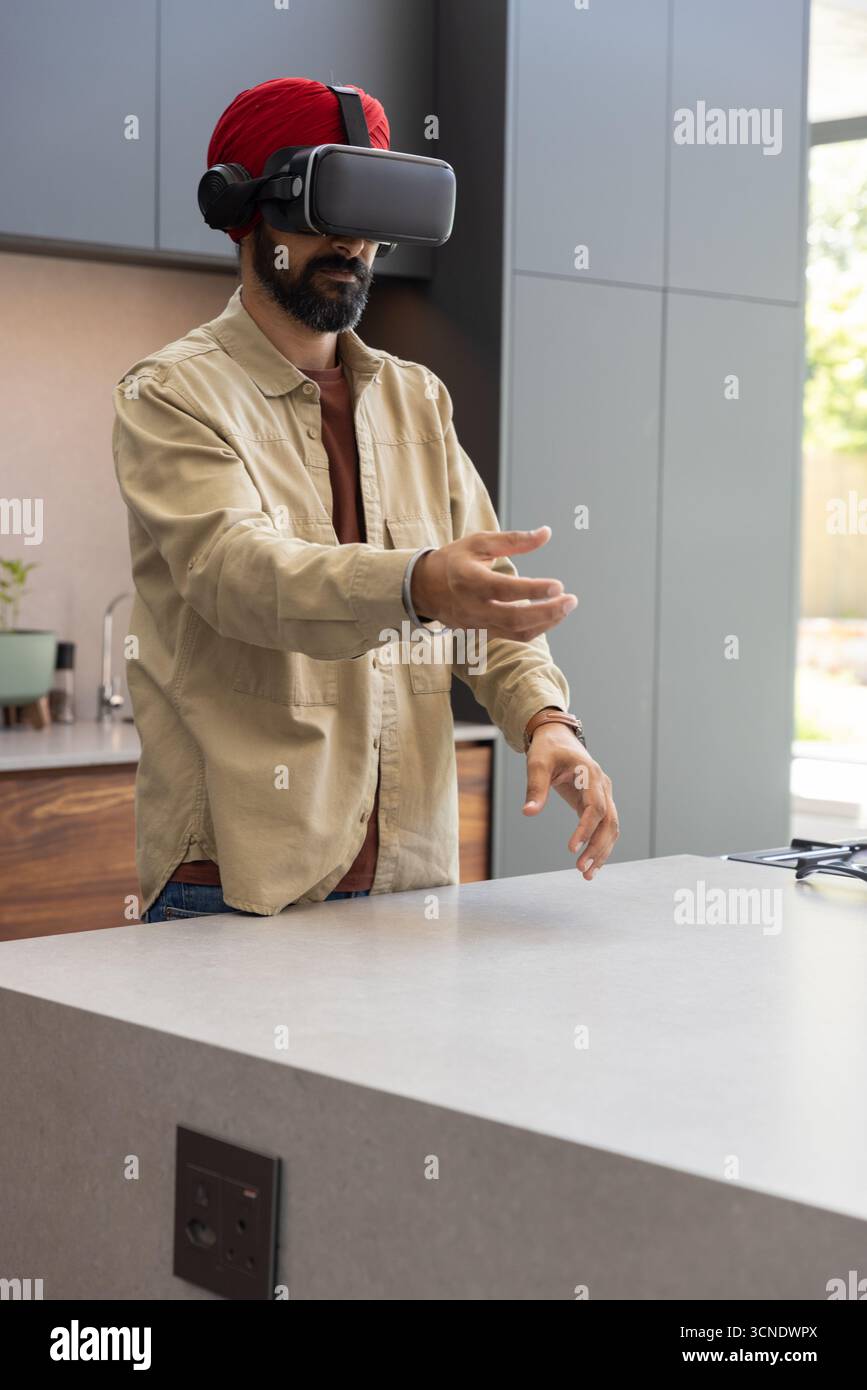 Indian man using vr headset with headphones at kitchen island, manipulating virtual interface Stock Photo