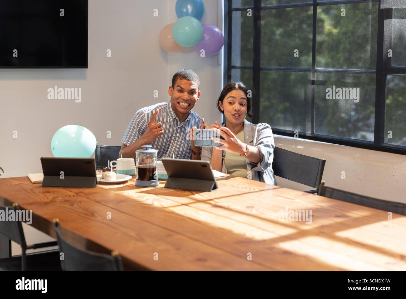 Diverse coworkers taking selfie with smartphone at meeting desk amid balloons, tablets, mugs Stock Photo