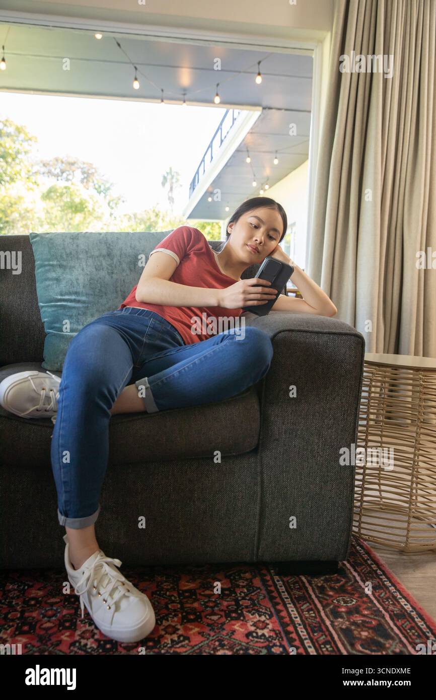 Asian woman reclining on gray couch in living room leaning against teal pillow, using smartphone Stock Photo