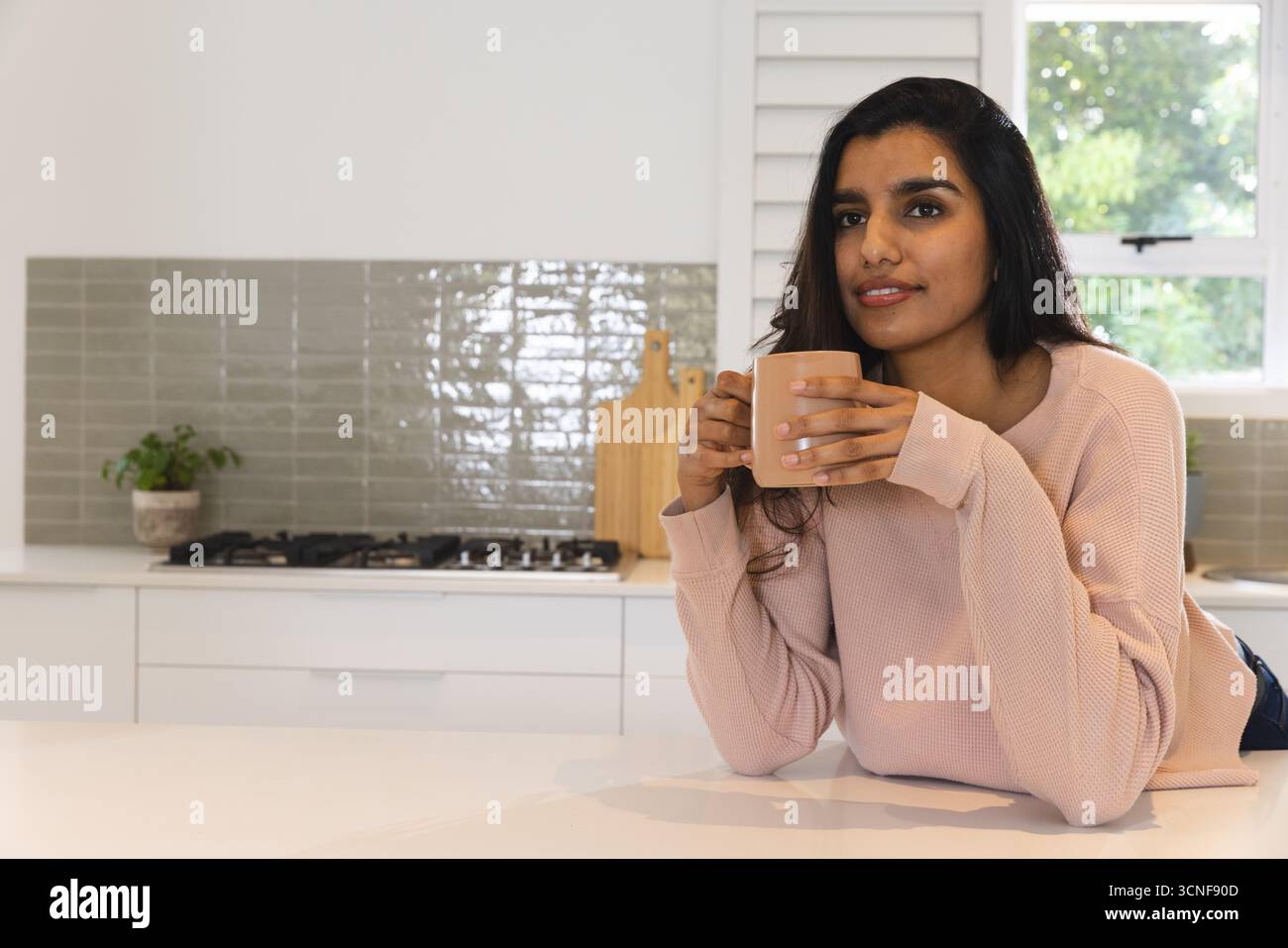 South Asian woman leaning on white kitchen island holding light brown ceramic mug, copy space Stock Photo