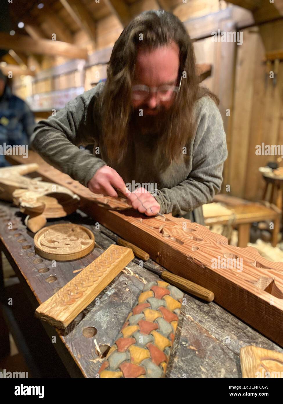 A Viking carpenter busy carving a long piece of wood to fit part of a warship in his workshop in the reconstruction of the Yggdrasil CBarn inside the - Smartphone Captured Stock Image